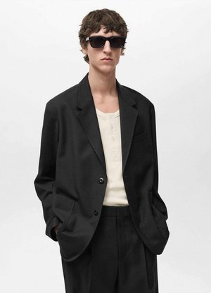 Blazer nero con una vestibilità sartoriale, abbinato a una maglietta henley crema. Il modello indossa occhiali da sole neri, mostrando un look minimalista.