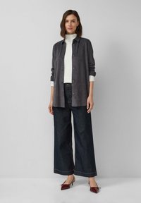 Graue Wildlederhemdjacke mit Knopfverschluss, getragen über einem weißen Rollkragenpullover. Kombiniert mit weit geschnittenen, dunklen Jeans und bordeauxfarbigen spitzen Absätzen.