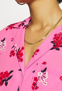 Camicia floreale rosa con fiori rossi e bianchi e foglie nere. Collo corto, taschino singolo e una texture di tessuto liscia. Collana in oro.