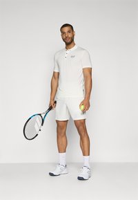 Λευκό πόλο μπλουζάκι με μικρό μπλε λογότυπο EA7, συνδυασμένο με άσπρα σορτς. Το άτομο κρατά μια ρακέτα τένις και μια πράσινη μπάλα τένις.