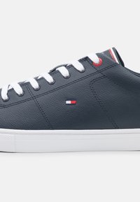 Marineblaue Sneaker mit weißer Sohle und Schnürsenkeln, kleinem rot-weiß-blauem Flaggenlogo an der Seite und rotem Akzent nahe dem oberen Ösenloch.