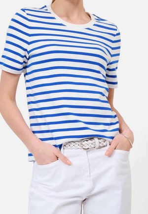 Nainen, jolla on valkoinen ja sininen vaakaraitainen lyhythihainen paita, valkoiset housut ja punottu valkoinen vyö, kädet taskuissa.