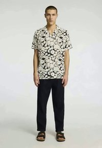 Camisa de manga curta com um padrão floral preto e branco, combinada com calças escuras e sandálias castanhas. Tecido suave, corte descontraído.