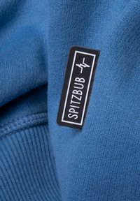 Blauer Pullover mit einer weichen Textur, versehen mit einem schwarzen Etikett, auf dem "SPITZBUB" in weißer Schrift steht. Sichtbare gerippte Saumdetails.