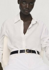 White button-up shirt made of lightweight fabric, worn over high-waisted white pants, featuring a black leather belt with a gold buckle.