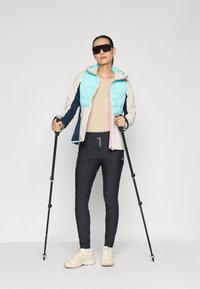 Lichte, veelkleurige jas met een turkooise geïsoleerde sectie, beige basis en marineblauwe mouwen; gecombineerd met zwarte cargo-broeken en beige schoenen.