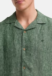 Camicia colllare in lino verde con una finitura testurizzata, caratterizzata da bottoni in legno e un piccolo logo ricamato sul petto.