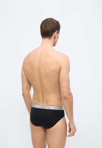 Οπίσθια όψη ενός άνδρα που φοράει μαύρα εσώρουχα με γκρι ζώνη μέσης που φέρει επαναλαμβανόμενο λογότυπο, στέκεται μπροστά σε απλό φωτεινό φόντο.