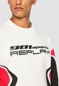 Hvit sweatshirt med fet svart tekst "981sports REPLAY." Røde og svarte detaljer på ermene gir et sporty design. Glatt tekstur.