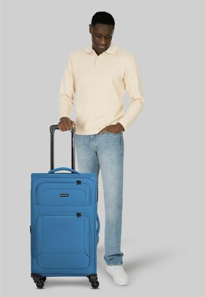 Man in beige lange mouwen shirt en spijkerbroek staat naast een blauwe koffer met wieltjes en een uitgeschoven handvat tegen een grijze achtergrond.