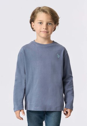 Camicia a maniche lunghe in cotone azzurro chiaro con colletto tondo e un piccolo logo ricamato sul petto, abbinata a jeans blu.