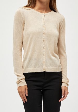 Mujer vestida con un cárdigan beige de manga larga abotonado y pantalones negros, de pie con los brazos relajados a los lados.