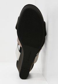 Semelle noire d'une sandale compensée Calvin Klein avec grip texturé et logo de la marque, montrant une partie des brides supérieures blanches et beiges.