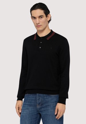 Schwarzer, langärmeliger Polo-Pullover mit einem kastanienbraunen Kragen, einer Drei-Knopf-Leiste und kleinem Logo auf der Brust; kombiniert mit blauen Jeans.