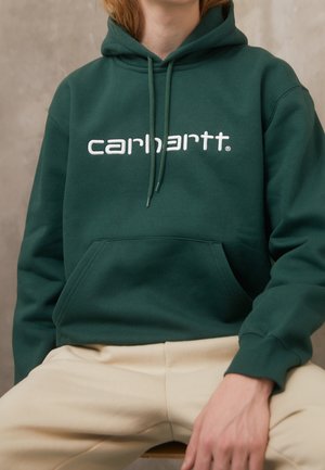 Personne portant un sweat à capuche Carhartt vert foncé avec un logo blanc et un pantalon beige, assise avec une main reposant sur le genou sur un fond neutre.