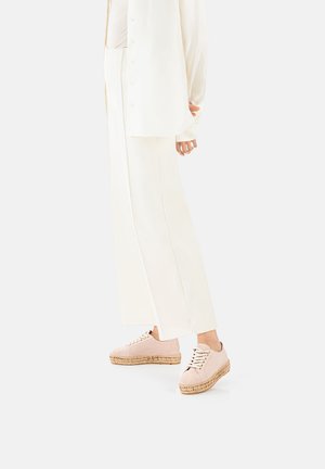 Light pink suede sneakers with woven jute sole, featuring rounded toe, lace-up design, and gold-tone eyelets, paired with white wide-leg pants.