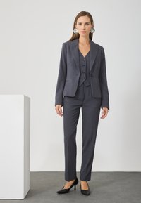 Fato cinza sob medida com um blazer ajustado, colete abotoado e calças de perna reta. Textura suave; saltos altos pretos combinando; brincos grandes.