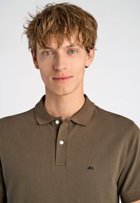 Ruskea polo-paita, joka on valmistettu tekstuurista kankaasta, jossa on kaulus ja kaksi nappia; rinnassa on pieni musta logo.