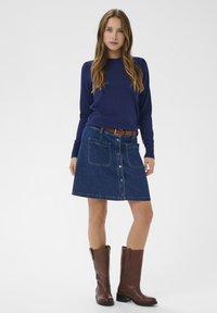 Pull en maille marine, jupe en denim bleu avec fermeture à boutons et poches avant, ceinture en cuir marron, et bottes en cuir marron jusqu'aux genoux.