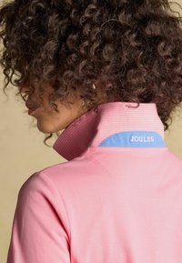 Kurruliste tumedate juustega isik, kes on küljele pööratud, kannab roosat kraega särk, mille tagumisel krael on sinine silt, millel on kiri "Joules".