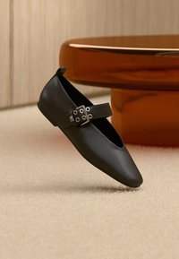 Μαύρο δερμάτινο επίπεδο παπούτσι με μυτερή μύτη και λουράκι με ασημένιες μεταλλικές λεπτομέρειες. Ομαλή υφή με μινιμαλιστικό σχεδιασμό.