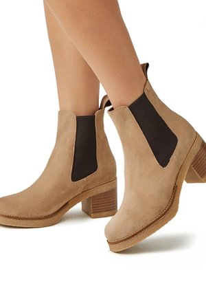 Botines de ante beige con lados elásticos negros, punta redonda y tacón de madera apilada. Suela de goma texturizada para mayor agarre.