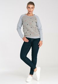 Jasnoszary sweter z teksturowanym przodem, kolorowymi kropkami i długimi rękawami. Noszony z ciemnymi dżinsami i białymi trampkami. Prosty, casualowy design.