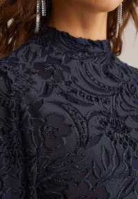 Marinblått broderat tyg har intrikata blommönster med texturvariationer. Hög krage med en genomskinlig finish.