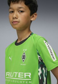 Jasnozielony sportowy strój z czarnymi akcentami, z logo, tekstem i wzorem kamuflażu na rękawach. Lekka tekstura i design.