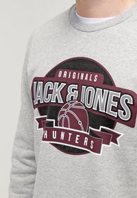 Sudadera gris con un gráfico audaz que presenta el texto "JACK & JONES" y un diseño de baloncesto, todo ello sobre un fondo circular burdeos.