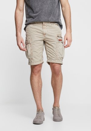 Mężczyzna ubrany w beżowe szorty cargo z kieszeniami, ciemnoszary t-shirt i szare sneakersy stoi na jednolitym tle.