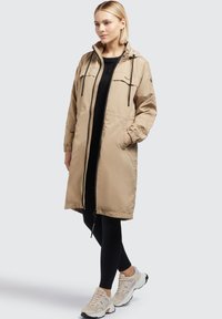 Beiger langer Parka mit Kapuze, Frontreißverschluss und zwei Seitentaschen. Verfügt über elastische Bündchen und eine lockere Passform, getragen über einem schwarzen Oberteil und Leggings.