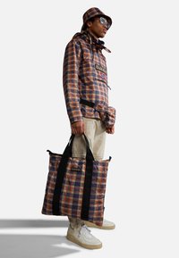 Uomo che indossa una giacca a quadri, un cappello a secchiello abbinato e un marsupio, pantaloni beige e sneakers bianche, mentre tiene una grande borsa a quadri con manici neri.