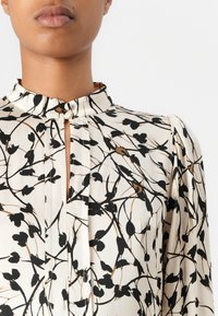 Cremefarbene Bluse mit schwarzem Blumenmuster, verfügt über einen hohen Kragen und eine kleine Schlüsselöffnungsdetail, gefertigt aus glattem, leichtem Stoff.