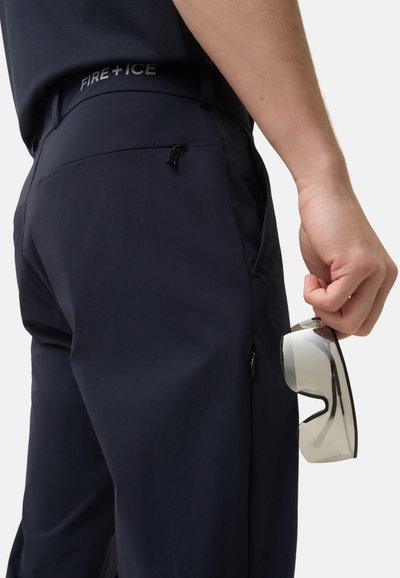 Námořnické modré kalhoty s elegantním designem, vybavené zipovou kapsou a reflexním logem na pase. Ruka drží průhledné sluneční brýle.