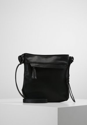 Sac bandoulière en cuir noir avec une sangle ajustable et une poche avant zippée, exposé sur un socle blanc sur un fond uni.