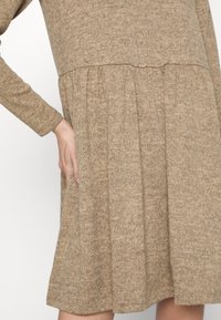 Ein beiges, strukturiertes Strickkleid mit langen Ärmeln, einem figurbetonten Oberteil und einem gerafften, ausgestellten Rock. Die Nahtdetails entlang der Taille sind sichtbar.