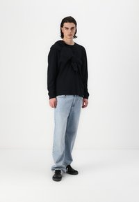 Melnas garām piedurknēm krekls ar sasietu džemperi pāri plecam, gaiši zili platās bikses un melni sporta apavi uz modeļa. Balta fonā.