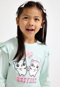 Licht mintgroen langarmig shirt met ruches, met een kleurrafische afbeelding van twee cartoonkatten en de tekst "BESTIES" in roze.