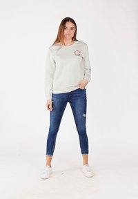 Felpa grigia chiara con grafica rotonda rossa, abbinata a jeans slim-fit blu e sneaker bianche. Texture liscia, design casual.