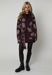 Vinröd stickad kofta med paisleymönster, fransiga kanter och matchande skärp. Bärs över en svart topp och kombineras med svarta knähöga stövlar.