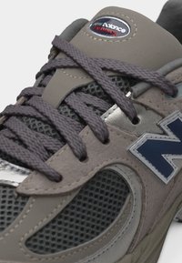 Κοντινό πλάνο γκρι και ναυτικού μπλε αθλητικού παπουτσιού New Balance με ύφασμα από δίχτυ, επενδύσεις σουέντ, χονδρές κορδόνια και λογότυπο της μάρκας στη γλώσσα και στην πλευρά.