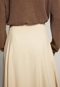 Pull en tricot marron à manches longues, rentré dans une jupe taille haute beige en ligne A, en tissu lisse avec des coutures discrètes.