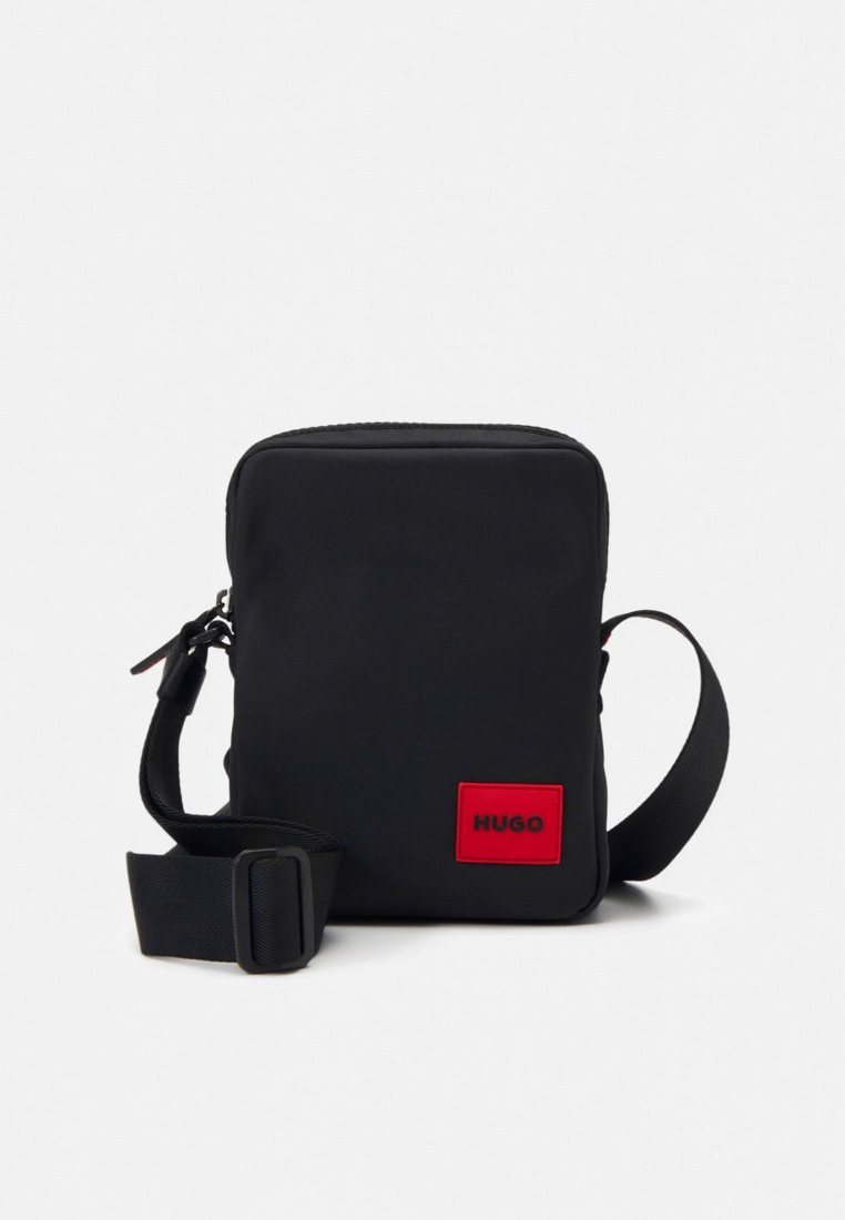 Sac bandoulière rectangulaire noir avec sangle réglable et patch carré rouge portant l'inscription "HUGO" à l'avant.