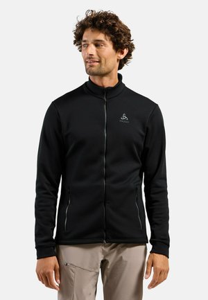 Homme aux cheveux bouclés portant une veste noire zippée et un pantalon beige, debout et regardant vers sa gauche sur un fond uni.
