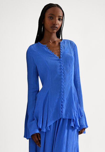 Naine pikkade patsidega, seljas tekstuurse lõiguga kobolt-sinise pikkade varrukatega pluus, millel on kangaskatetega nupud ja lehvikukujulised mansetid, taustaks lihtne taust.