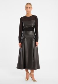 Schwarzes Lederkleid mit langen transparenten Ärmeln, tailliertem Oberteil, ausgestelltem Rock mit Taschenakzenten, kombiniert mit goldenen metallischen High Heels.