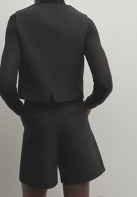 Gilet noir ajusté avec un col haut et des manches courtes, associé à un short tailleur noir, doté de poches latérales et d'une texture de tissu lisse.