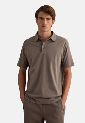 Joven con cabello castaño que lleva una camiseta polo marrón de manga corta y pantalones a juego, de pie con las manos en los bolsillos.