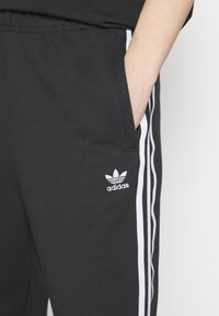 Černé tepláky vyrobené z bavlny, s bílými bočními pruhy, elastickým pasem a malým bílým logem Adidas na levém boku.